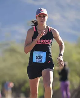 A person running in a race, wearing a black tank top with "ROAD ROCK" written on it, black shorts, and a visor. They have a race bib numbered 535. The background shows a blurred outdoor setting of trees and hills.